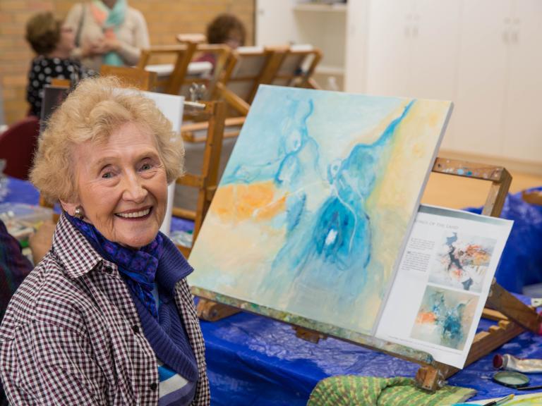 An older woman smiles at the camera with her painting sitting behind her