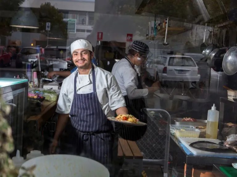 A chef standing in a busy kitchen wearing white clothes and a blue apron, and holding a plate of pasta