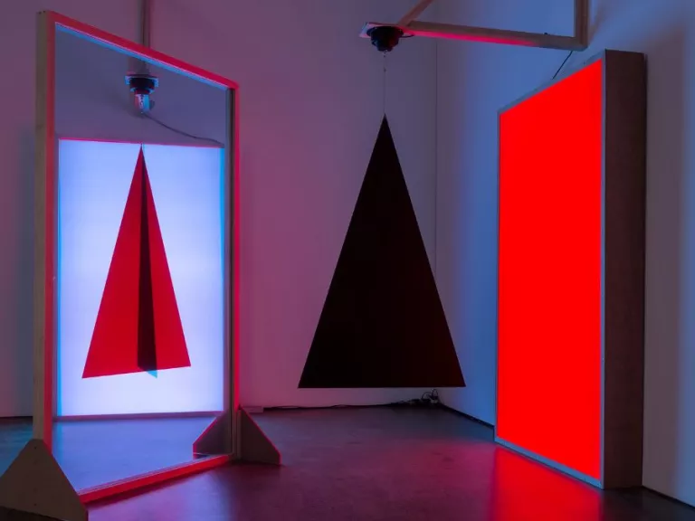 A mirror reflecting the image of a red triagle with a black line through the centre, next to a bright red lit rectange and a hanging 3D black pyramid in front of it.