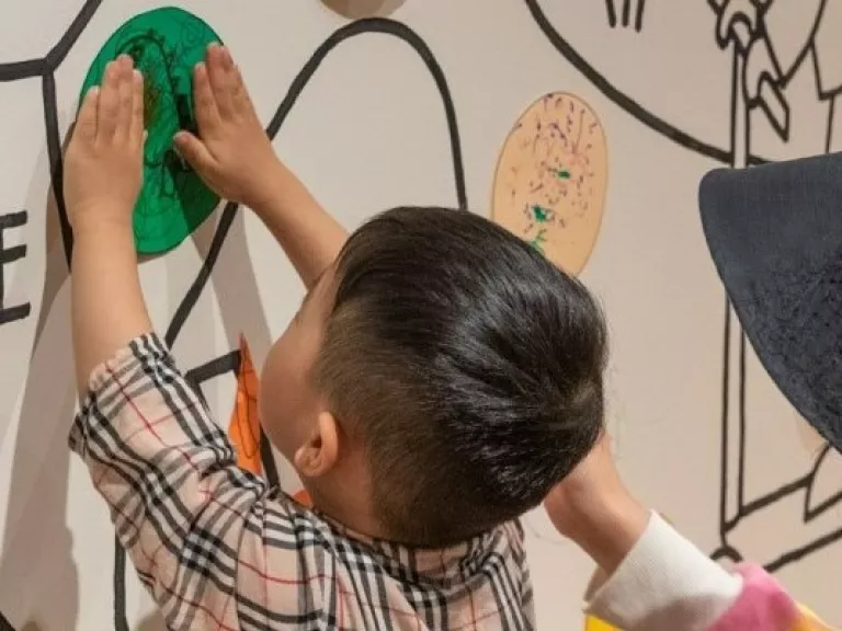 A young child with an adult, adding a piece of craft to a mural on a wall