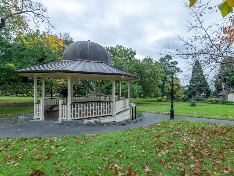 Rotunda with cream-white balustrades and grey dome-shaped roof, surrounded by a footpath and grass area. There is a lamp post in the centre and fallen leaves on the ground.