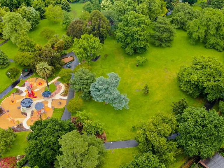 Aerial view of grass area with many scattered green trees. There is a large playground with surrounding footpaths to the left.