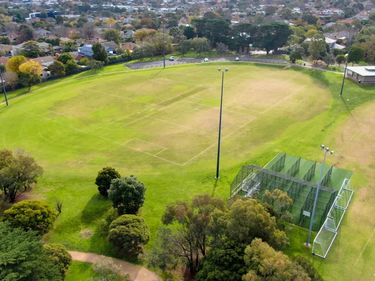 Aerial view of oval sportsground with various square line markings on its surface and four tall light posts around its edge. There is a pavilion to the top right and cricket nets to the bottom right.