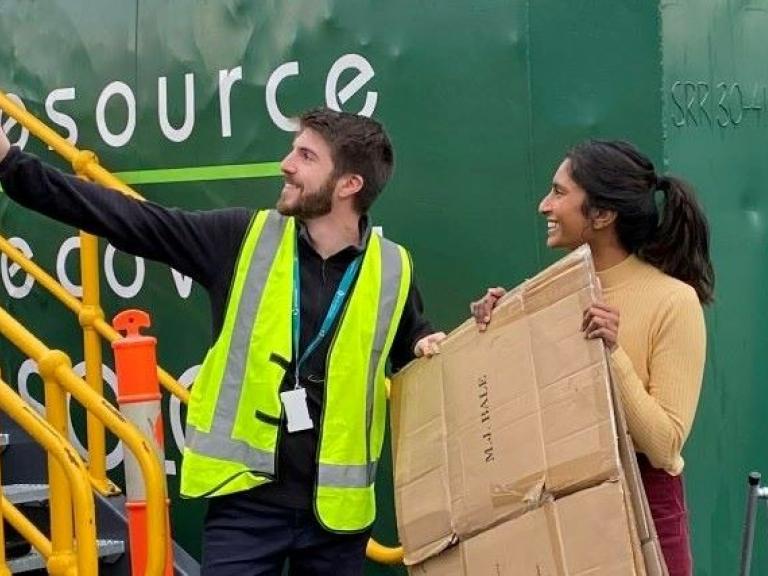 A person holding a flattened cardboard box being directed by a staff member where to put it at the recycling depot