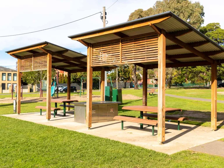 Barbecue area in a park, with 2 sets of tables and chairs and 2 wooden shelter structures with diagonal roofing.