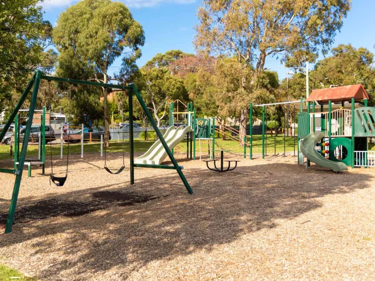 Playground on a mulch surface. It has swings and two climbing features with slides, surrounded by a grass area and tall trees.
