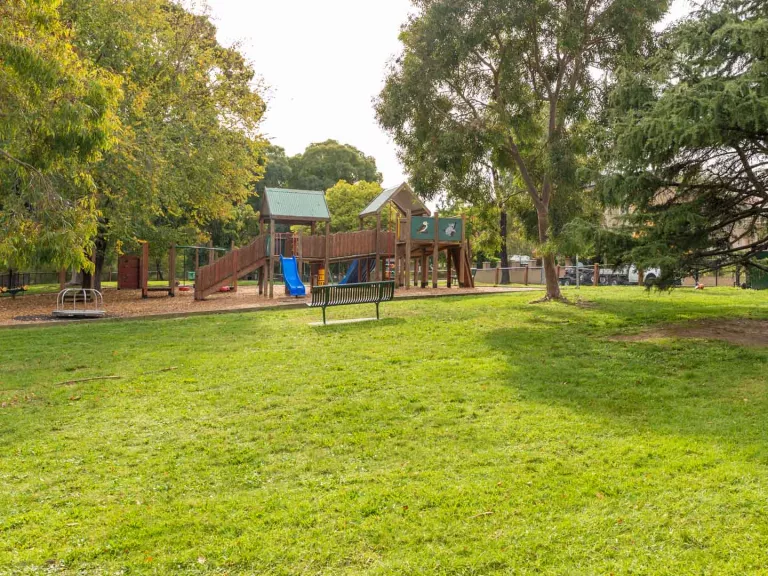 Small grass area with park bench facing a playground, surrounded by scattered tall trees.