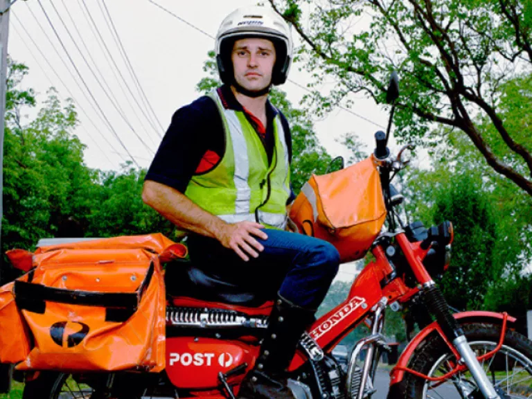 Photograph of a postman on a red Australia Post motorcycle. Orange mailbags are attached to the bike, and the postman is wearing a high-vis vest and white helmet. He is stopped in the middle of the street with one foot on the ground to steady the bike looking directly at the camera. The background shows a tree-lined street with wheely bins out for collection.