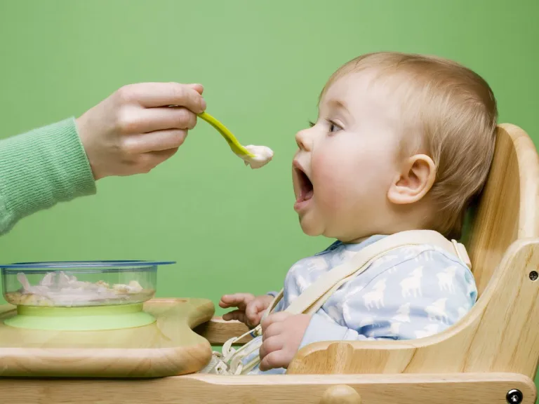 baby in high chair being fed food from a person off screen with solid food in bowl on highchair 