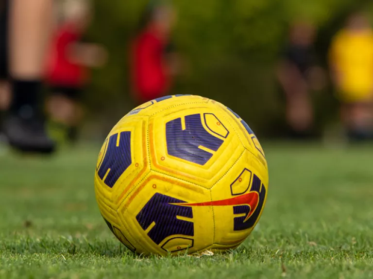 A black and yellow soccer ball sits on grass with several players in the background.