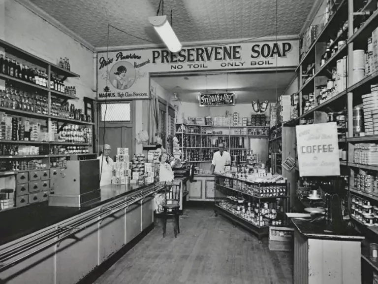 Black and white photograph of a grocer shop. Shelves lined with tinned and bottled goods line the walls of the store and there is a long table with a cash register. Three staff are in the background, one behind a counter at the back of the shop and two near the register partially concealed by product displays.