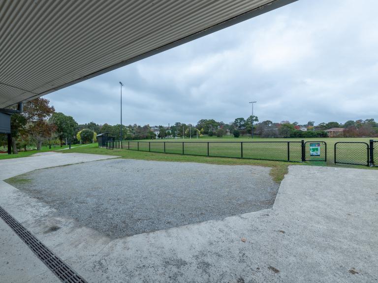 A view of the fenced sportsground. Surrounding the field is a concrete and gravel area under a roofed area. 