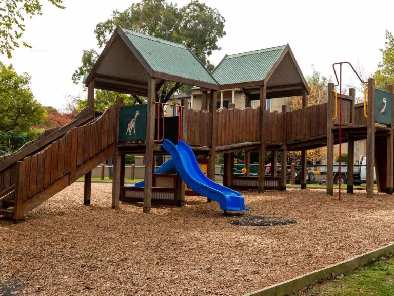 A wooden playground on a mulch surface. The playground ahs a slide and a ramp. 
