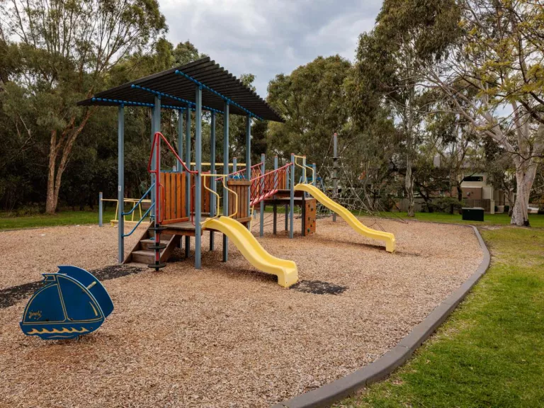 A playground with a bark flow. It has 2 slides and a rocking seat. A barbecue can also been seen.