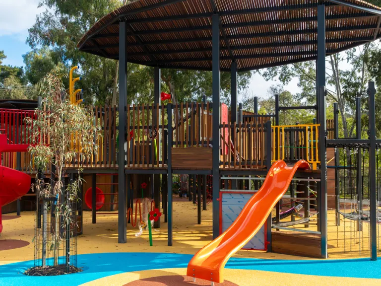 A multi-level play structure with a wooden base and a black metal roof. The structure includes two slides, one red and one orange, as well as a variety of climbing components and climbing walls. The play area is surrounded by trees and shrubs, and the ground is covered with rubber mulch in a variety of colors.