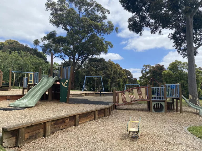 A playground with slide, climbing equipment and large trees providing shade