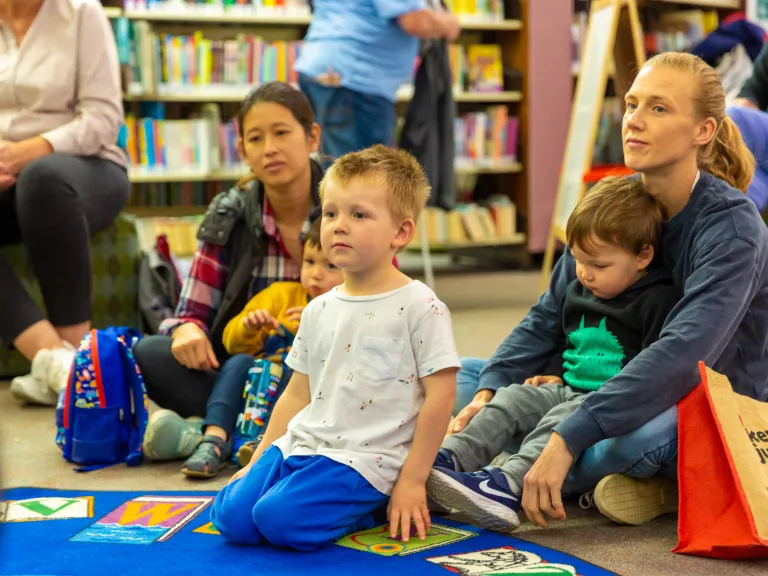 A group of children and their carers sit on a blue lap in a library.