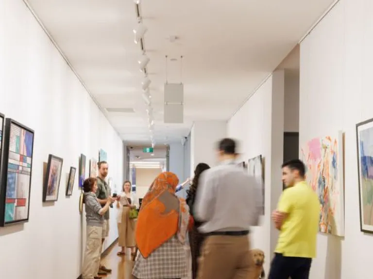 The image shows a long, well-lit gallery space with wooden flooring and white walls adorned with various artworks. The artworks include colourful abstract paintings, geometric compositions, and landscape scenes, all framed or mounted on the walls. Visitors are walking through the space, some engaged in conversation while others observe the pieces. Track lighting runs along the ceiling, illuminating the artworks. The perspective extends down the hallway.