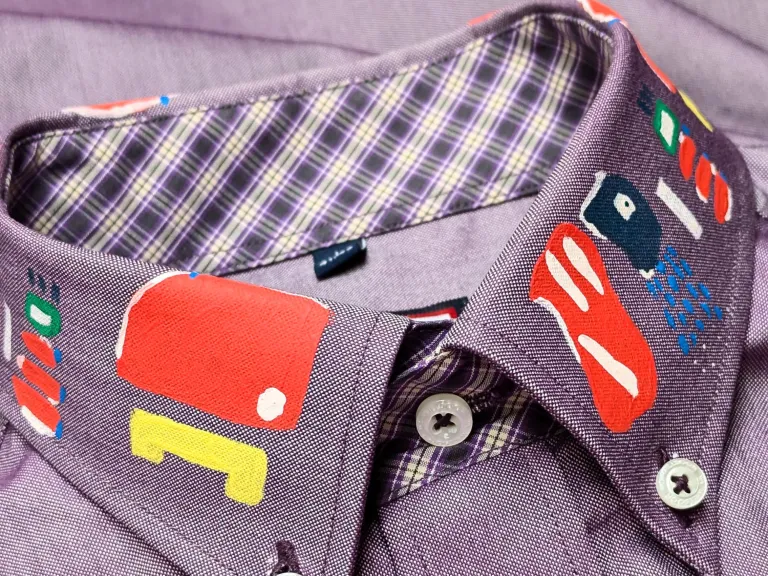 Close-up photograph of a buttoned-up purple shirt. The collar has been embellished with red, yellow, blue, green and white graphic shapes and patterns. 