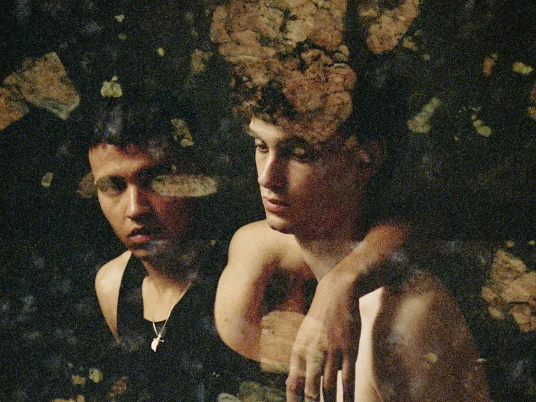 A photograph of two male figures sitting side by side against a black background. One has his arm over the shoulder of the other and is looking at him. The other is looking downwards and is holding a pale pink rose. Patches of semi-transparent brown botanical pattern cover the photograph. 