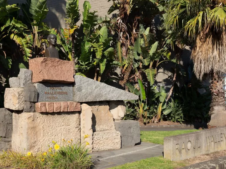 The sun shines brightly on a park with palms trees. In the foreground a sculpture stands proudly. A small bronze bust of a figure sits atop a large pile of angular stones
