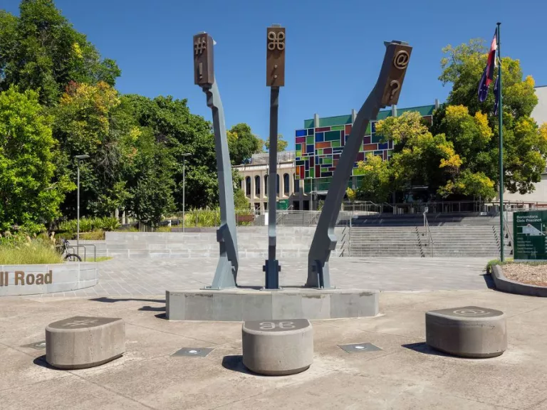 a monumental sculptures stands in a grey forecourt. Three arms that mimic the arms of a typewriter are reaching towards the sky. On the ground are three stones for the arms to land on