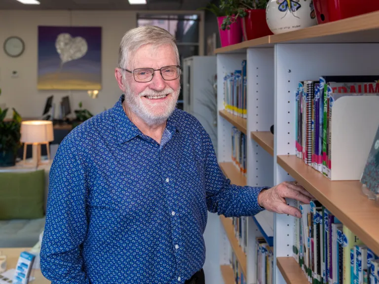 grey haired man wearing a blue shirt, standing at a bookcase