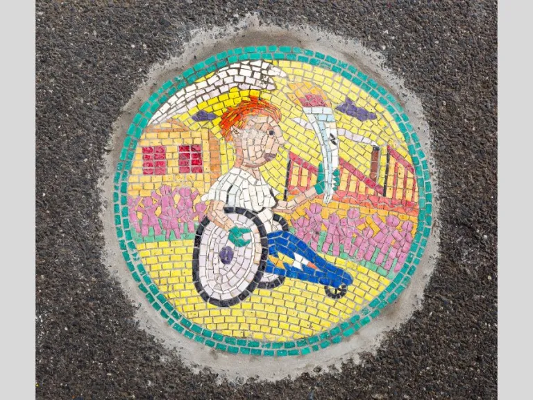 Small tile mosaic embedded in footpath depicting a figure in a wheelchair holding a flaming torch
