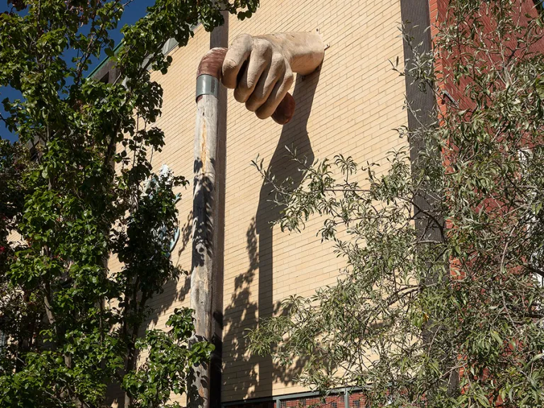 Sculpture of a large, fibreglass hand hand protruding from a brick wall and holding a wooden walking stick and surrounded by greenery.