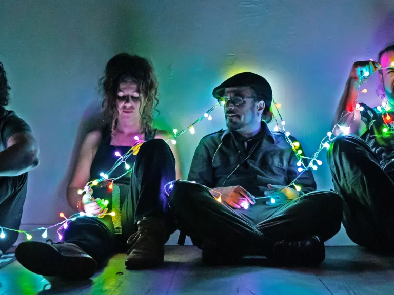 The four members of Bashka sit together in a house, a string of colourful fairy lights connecting them..