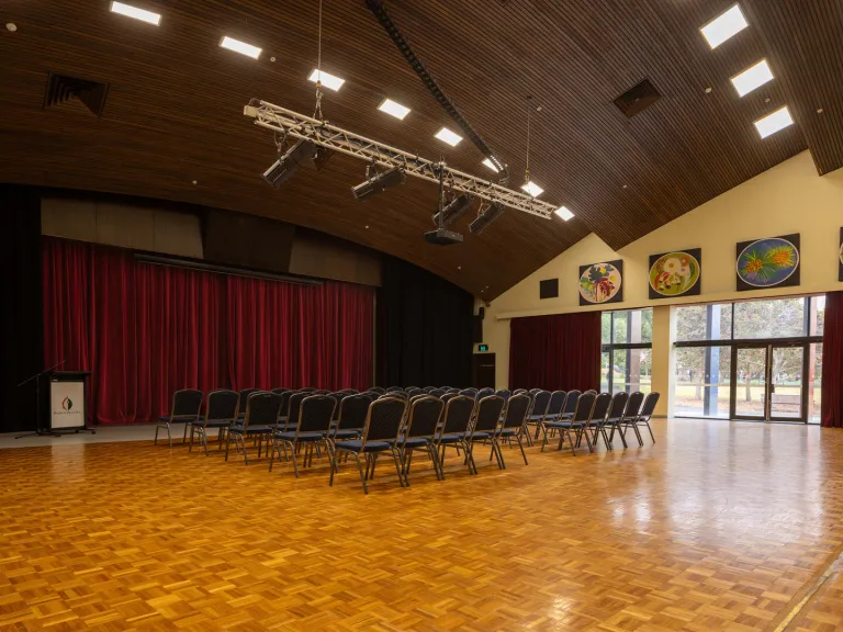 A room with rows of chairs facing a stage area with red curtains. Glass doors look out into a green area. The room has a wooden floor