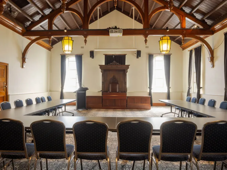 A conference room with long tables arranged in a U-shape, facing a raised platform with a wooden podium and antique-style furniture. The room has high, wood-beamed ceilings and arched details.