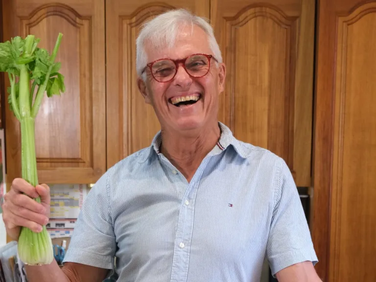 A middle-aged man in a wooden kitchen holds a stalk of celery and beams