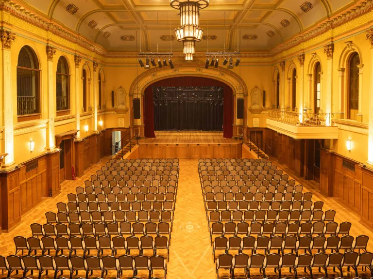 The spacious auditorium's interior includes a stage. Rows of chairs are set up in the main floor. There is a balcony on the right side and a large stage with red curtains at the far end. The room has ornate architectural details and is lit with chandeliers.