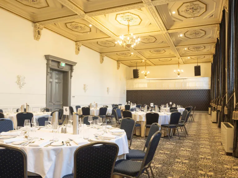 A large room is set up for a formal dinner. Round tables with white tablecloths are arranged throughout the room, each set with silverware, plates, and glassware. Blue chairs with a gold polka dot pattern are around the tables. The room has high ceilings with decorative molding, ornate panels and several chandeliers. A raised stage area with speakers is in the background. Dark curtains are on the right. The floor has floral pattern carpet.