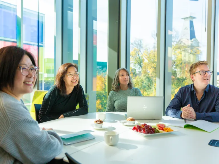 “Four people sit around an office desk during a meeting. The desk holds notepads, a laptop, and a plate of fruit.