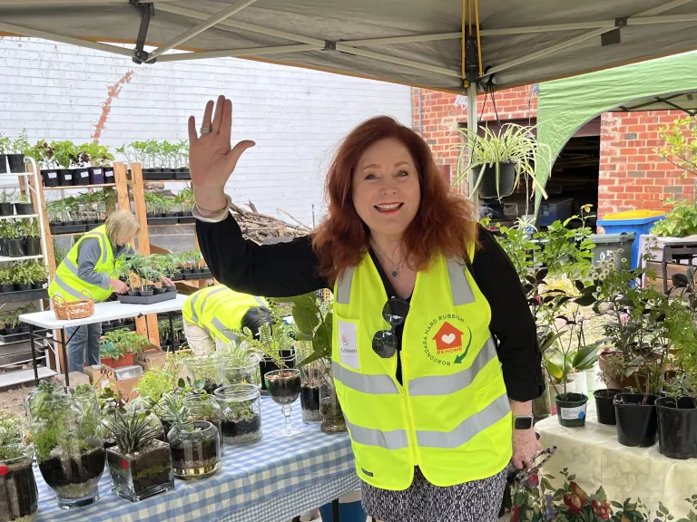 person standing in front of a plant stall at a market, smiling and waving. They are wearing a fluro yellow vest that says 'Boroondara Hard Rubbish Rehome'