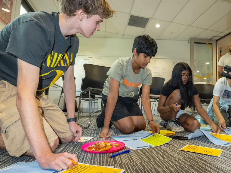 A group of young people sit on the floor of an office space. They are in a workshop.