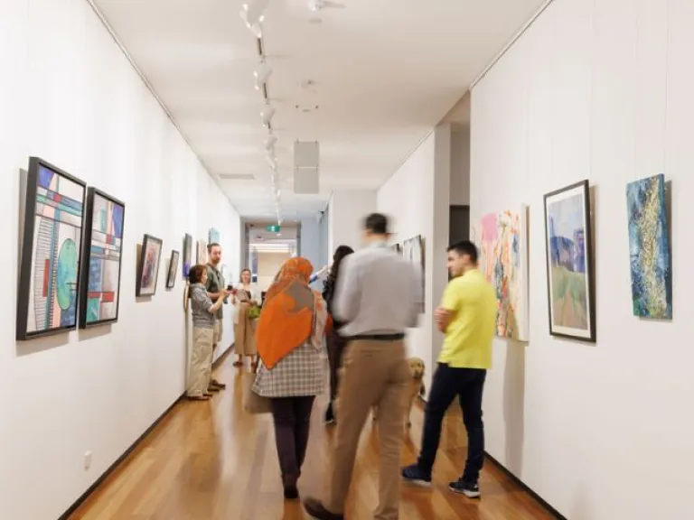 The image features a gallery hallway with paintings hanging on both walls. The artworks vary in style and colour, including landscapes, abstract pieces and geometric shapes. Several people are standing or walking through the space. 