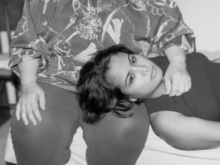 A black and white photo portrait of a young person lying with their head on an older person’s lap. The older person sits upright, hands resting on their legs, while the younger person looks directly at the camera.
