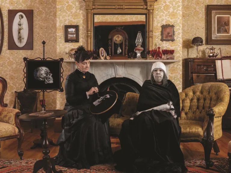 A staged Victorian-style interior with patterned wallpaper and antique furniture, where two women (artists megan evans and Maree Clarke) dressed in black sit together on an upholstered chair.