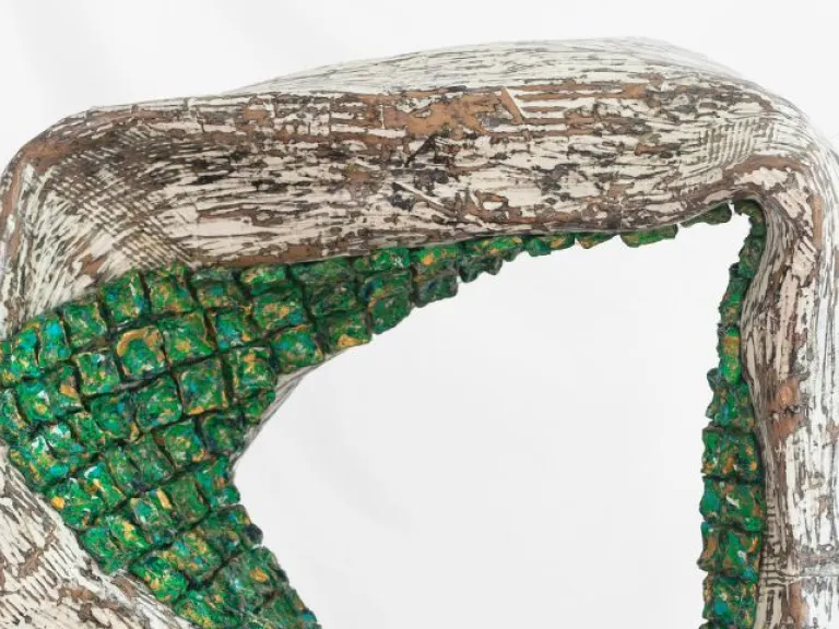 Close-up of a sculptural form with a rough, textured surface in white and brown tones. An arched opening is filled with small, clustered green blocks that resemble carved or moulded cubes. The overall shape curves organically, with etched, scratch-like markings across its surface.
