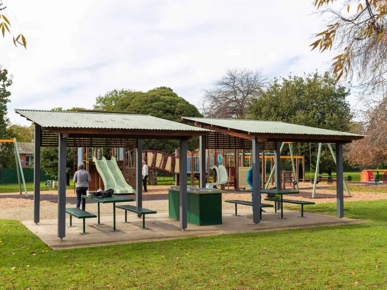 2 sheltered picnic shelters with a barbeque in between. the background is a children's playground