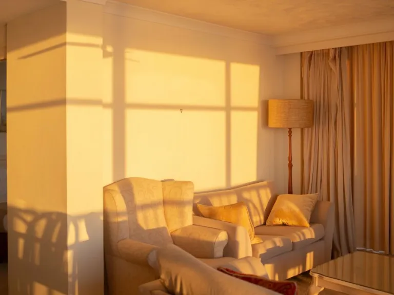 A sunlit living room including comfy chairs, a lamp, curtains and a hook on the wall that is missing an artwork. 