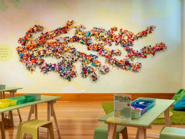 Wide gallery installation view showing a large wall artwork made from dense clusters of brightly coloured fabric flowers arranged in branching forms, with pastel-green worktables, trays of materials, stools, a rug and beanbags.