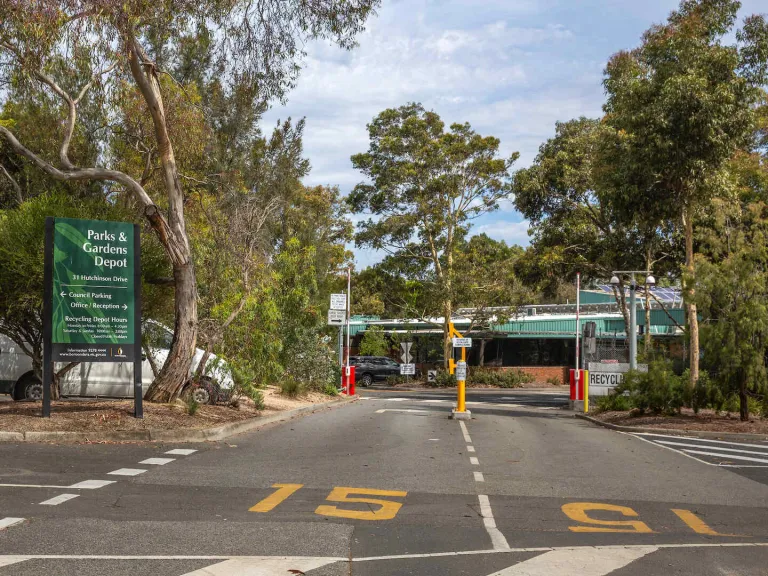 Paved entrance to the Parks and Gardens Depot, with a large green sign on the left, a boom gate down the centre road, and leafy trees surrounding the area.