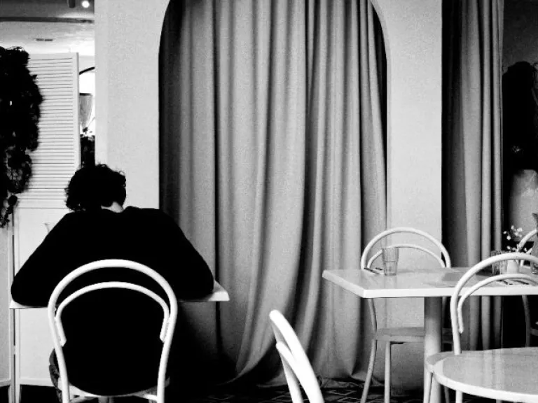 A black-and-white photo of a person seated alone at a table in a cafe. They are facing away from the camera. The space is filled with white chairs and tables, and an arched curtain feature dominates the background. Indoor plants add contrast to the scene.