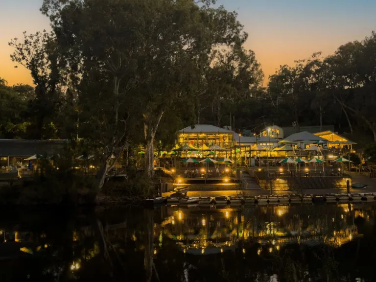 Photograph of a multi-story building lit with warm lights and surrounded by green and white umbrellas by a river at dusk. The building is surrounded by tall trees, and the sky is orange and blue. The lights of the building are reflected clearly in the still water below.