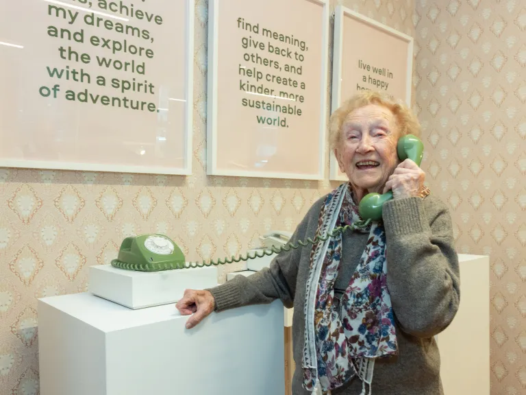Woman holing a corded phone handpiece to her ear. the phone is green, and placed against a cream patterned wallpaper. On the wall are 3 frames with text.