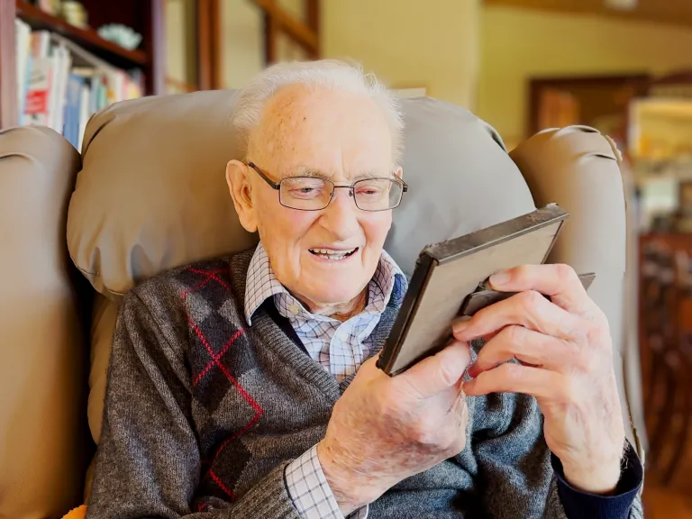 A balding man with white hair and glasses sits in a comfy armchair. He's looking fondly at a picture frame in his hands.